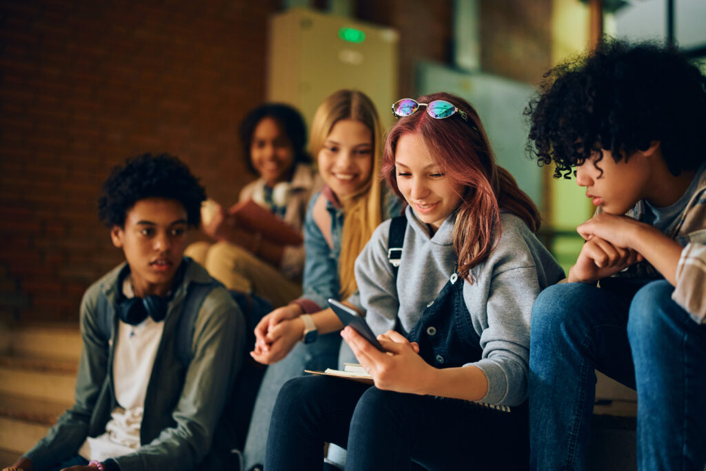 Happy high school student using smart phone with her friends in hallway.