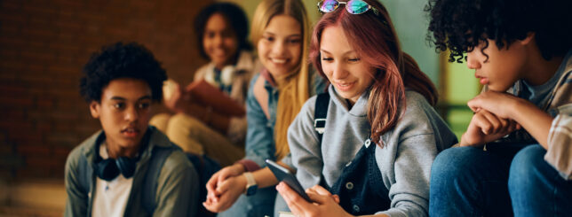 Happy high school student using smart phone with her friends in hallway.